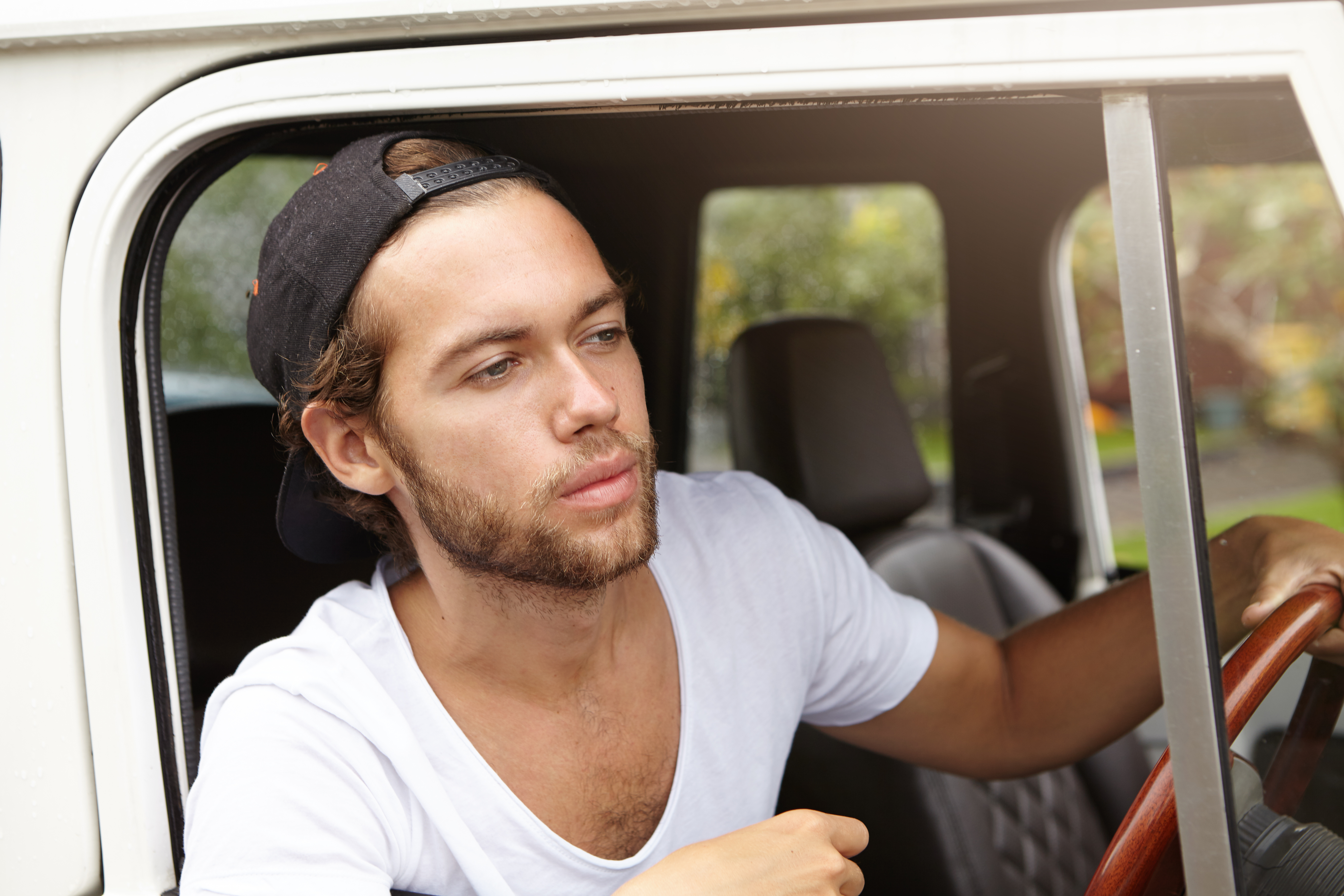 Young man in a white shirt and backward cap driving a vehicle, representing a baker delivering bagels with a thoughtful expression