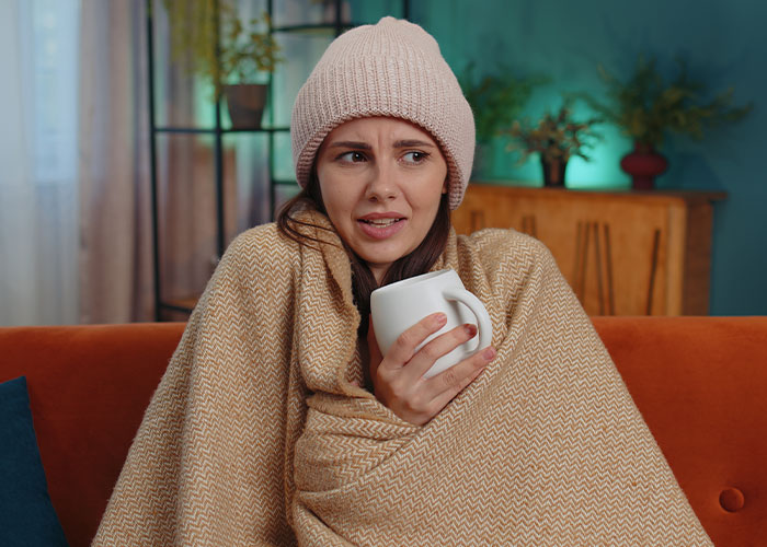 Woman wrapped in a blanket and wearing a hat, holding a mug and looking skeptical in a cozy indoor setting.