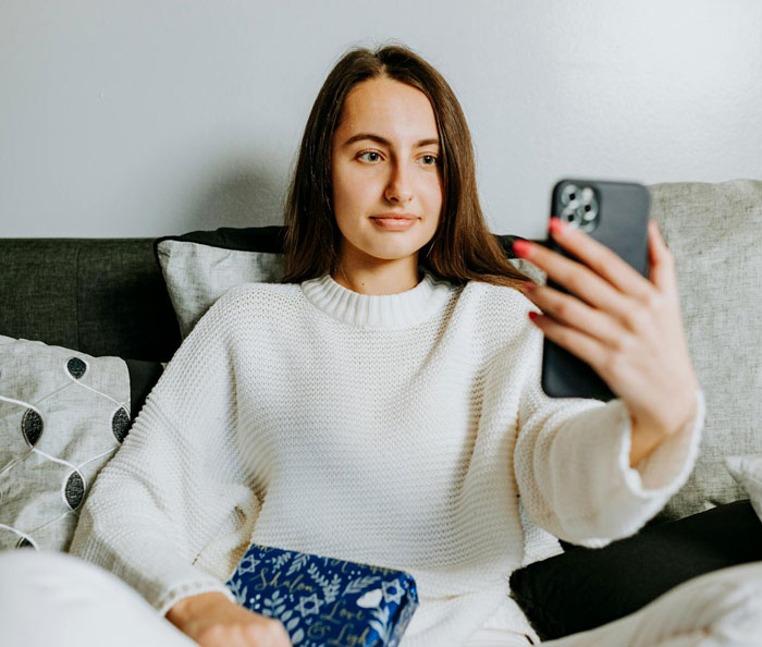 Young woman babysitting overnight, sitting on couch and holding phone, capturing a selfie with a calm expression.