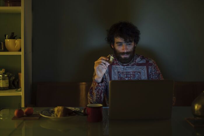 Man with beard sitting in the dark, looking at his laptop and eating, illustrating internet rabbit holes taking over lives.