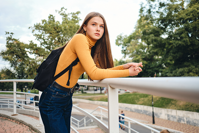 Teen girl in a yellow sweater and backpack leaning on railing, representing 17YO calling out mom retiring from parenting.