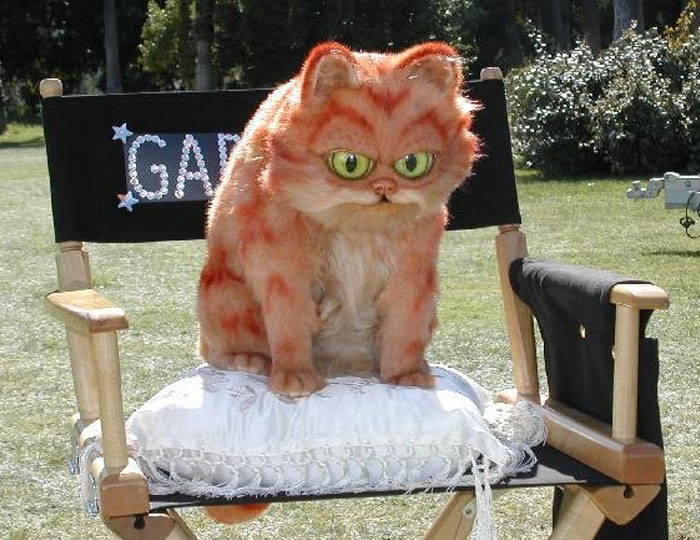 Unhinged behind-the-scenes photo of a large orange cat puppet sitting on a director’s chair outdoors.