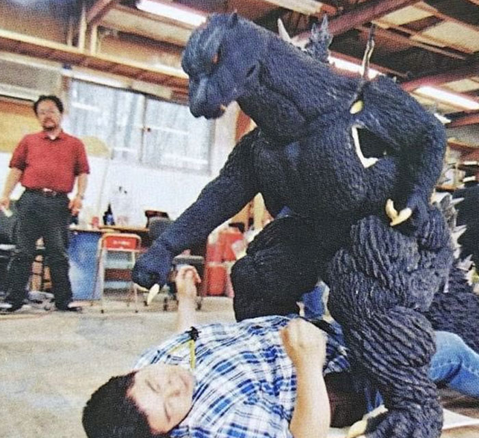 Unhinged behind-the-scenes photo of a person in Godzilla costume playfully interacting with a man lying on the floor in a workshop.