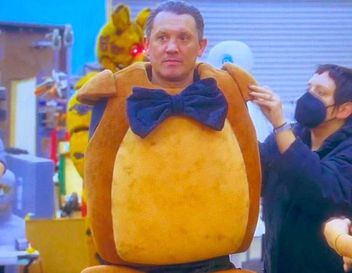 Actor in an unhinged behind-the-scenes photo wearing a large bear costume with a black bow tie being adjusted by crew.