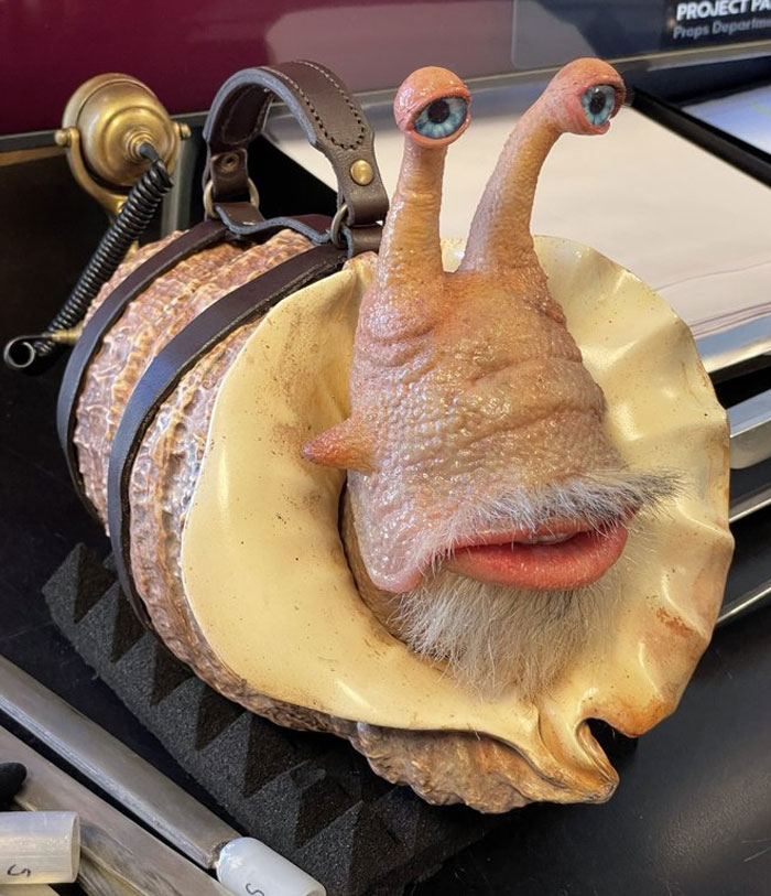 Unhinged behind-the-scenes photo of a realistic snail puppet with human-like eyes, lips, and a mustache on a film set.