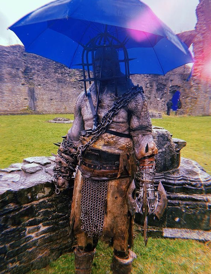 Unhinged behind-the-scenes photo of a medieval warrior costume with chains and an umbrella on a rainy day outdoors.