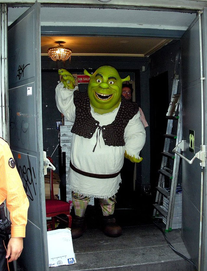 Person dressed as Shrek in costume, standing behind-the-scenes in a dimly lit room with open doors.