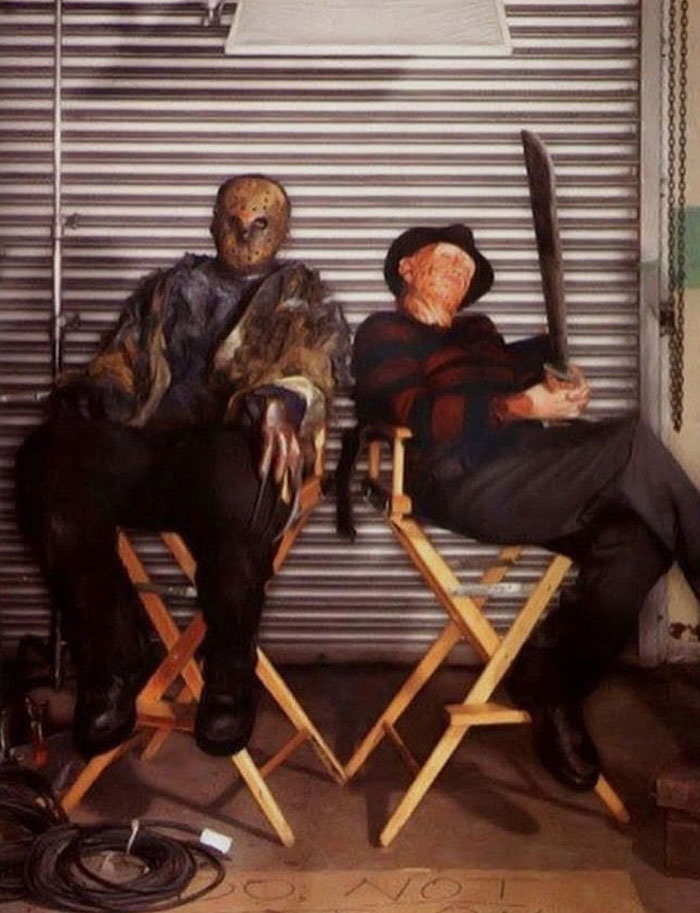 Two horror characters in costume sitting on director chairs in an unhinged behind-the-scenes photo setting.
