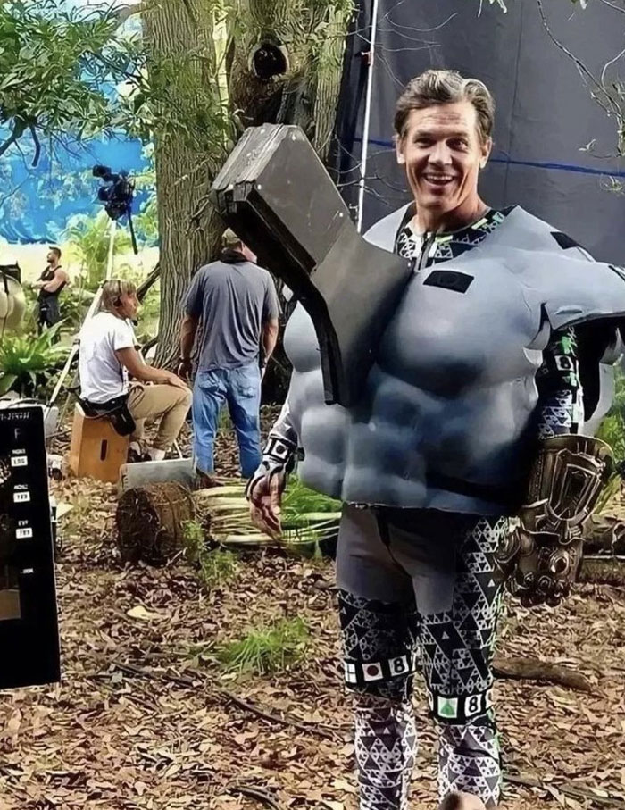 Actor in an unhinged behind-the-scenes costume with oversized chest piece on a forest set during filming.