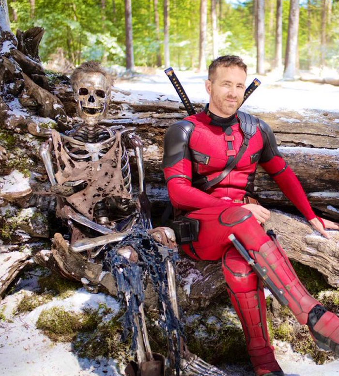 Deadpool actor in red suit posing with a skeleton prop in an unhinged behind-the-scenes photo outdoors.