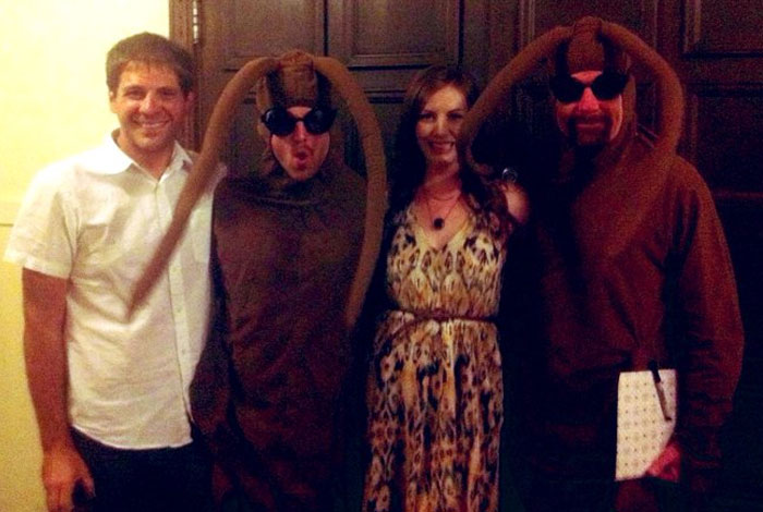 Four people posing for an unhinged behind-the-scenes photo, two dressed in brown cockroach costumes indoors.
