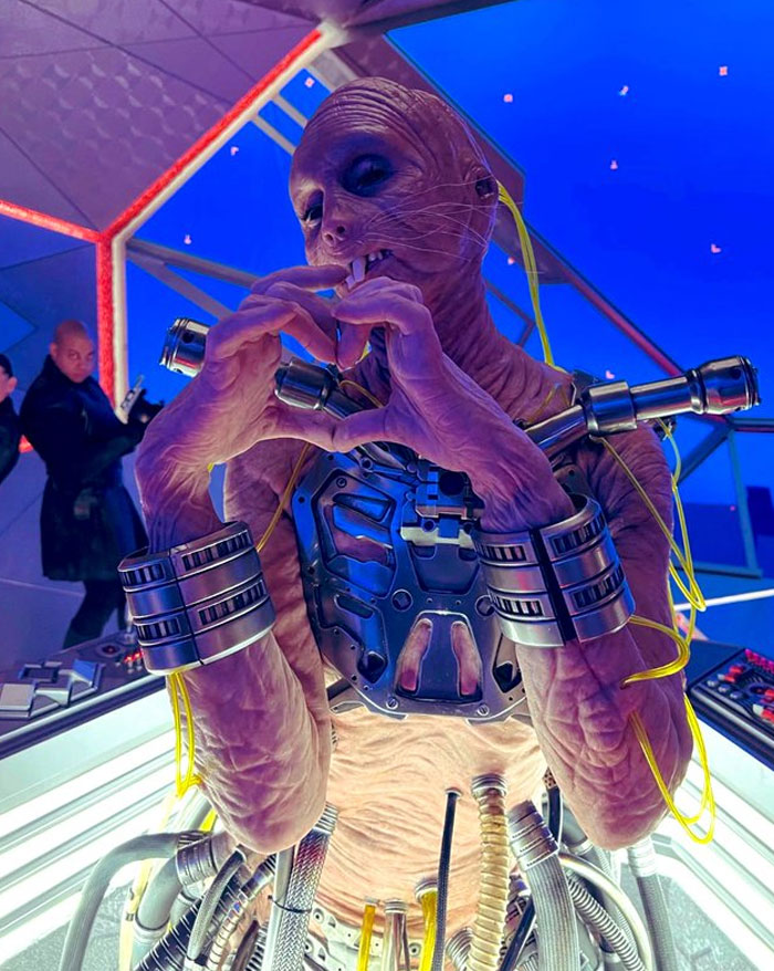 Unhinged behind-the-scenes photo of a sci-fi creature making a heart shape with hands surrounded by futuristic equipment.