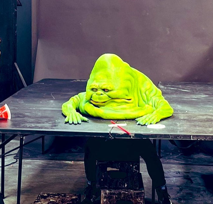Unhinged behind-the-scenes photo of a person in a green slime costume sitting under a black table in a studio.