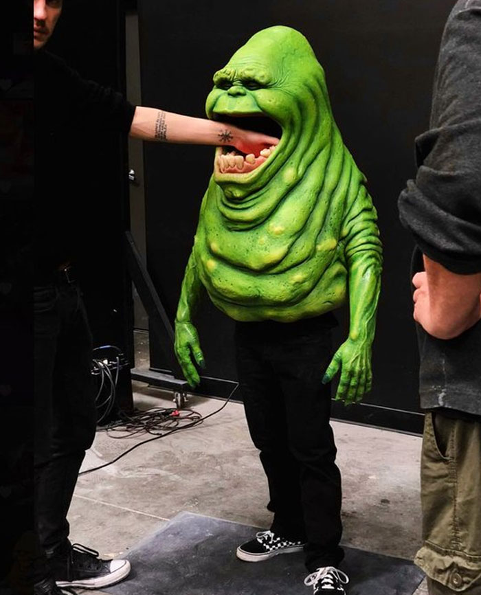 Behind-the-scenes photo of a person in a green creature costume with an arm playfully inside its mouth, meme moment.