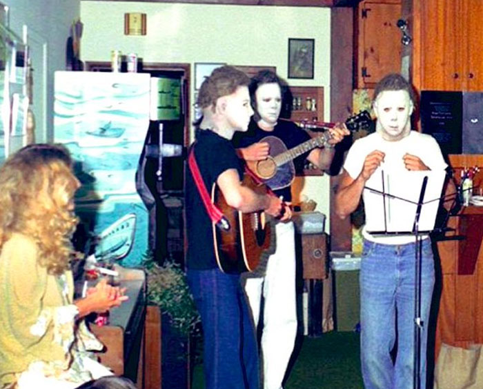 Three people wearing white masks play guitars and sing in a living room, an unhinged behind-the-scenes photo meme moment.