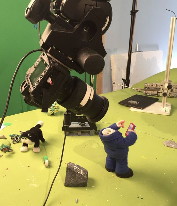Behind-the-scenes photo showing a camera filming a stop-motion puppet and toy cat on a green set for meme creation.