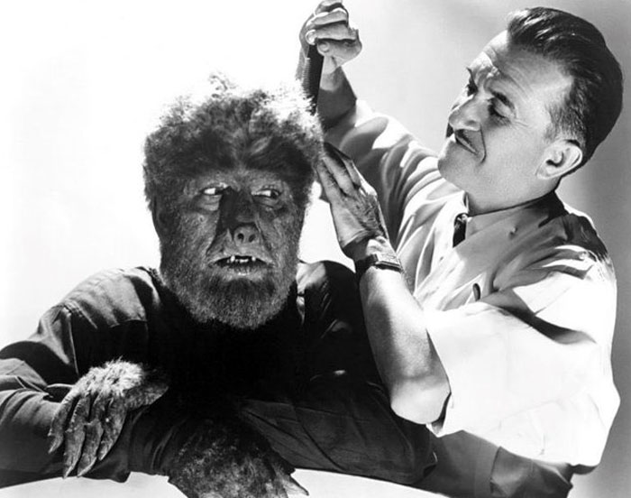 Behind-the-scenes photo of a man grooming an actor in detailed werewolf makeup for an unhinged movie scene.