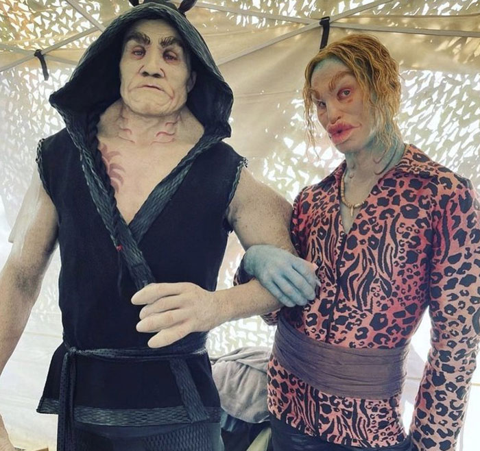 Unhinged behind-the-scenes photo of two actors in elaborate creature costumes posing inside a tent on set.