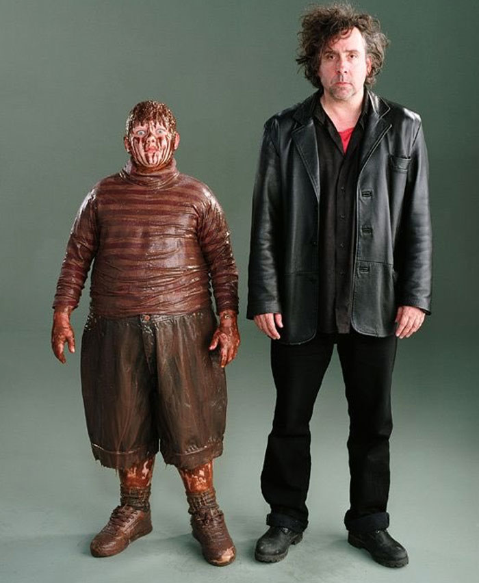 Two men in behind-the-scenes photo, one covered in fake blood, both posing against a plain gray backdrop.