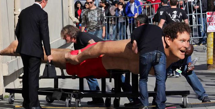 Large unhinged behind-the-scenes photo of a giant figure being transported by crew in a crowded outdoor area.
