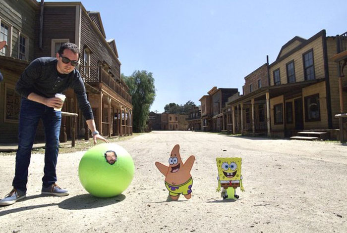 Man poses with green ball showing face behind-the-scenes with cartoon SpongeBob and Patrick cutouts on dusty street.