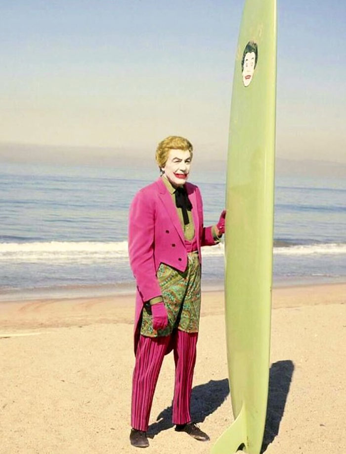 Person dressed as Joker in colorful outfit holding a surfboard on a beach, an unhinged behind-the-scenes photo meme moment.