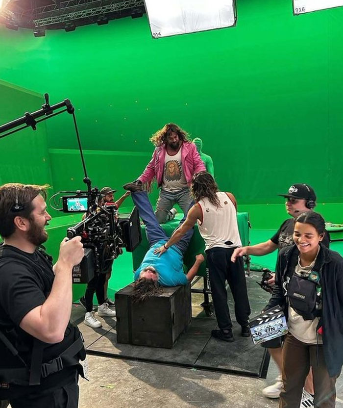 Behind-the-scenes photo of actors and crew filming a humorous scene on a green screen set with camera and crew members.