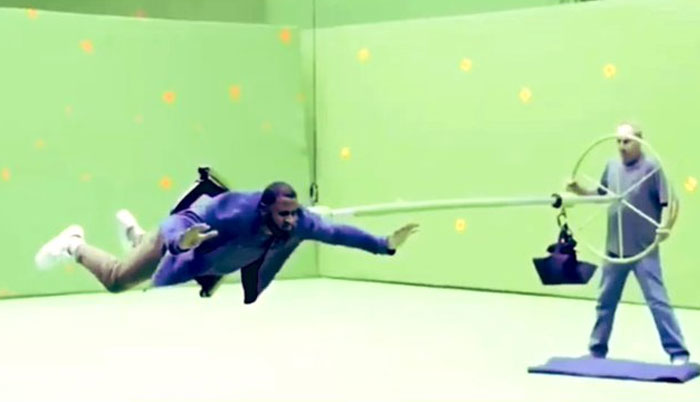 Actor suspended midair on wires in a green screen studio during unhinged behind-the-scenes photo shoot for memes.