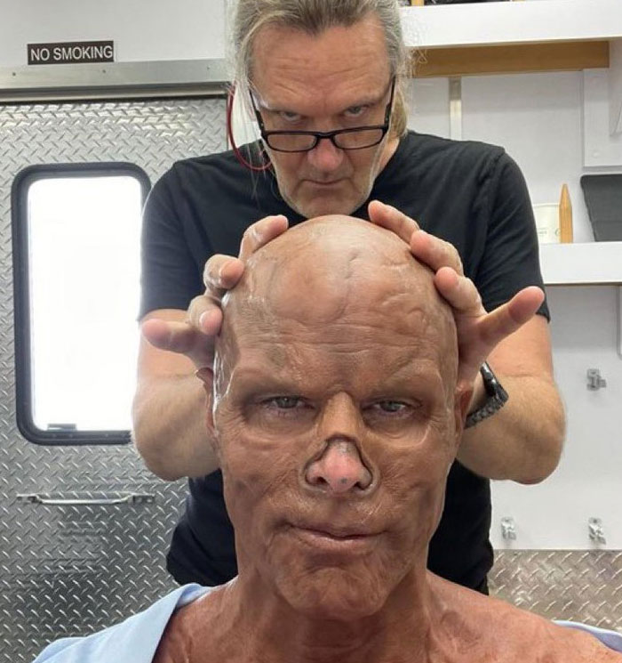 Man applying realistic mask in unhinged behind-the-scenes photo with intense expression in a makeup trailer setting.