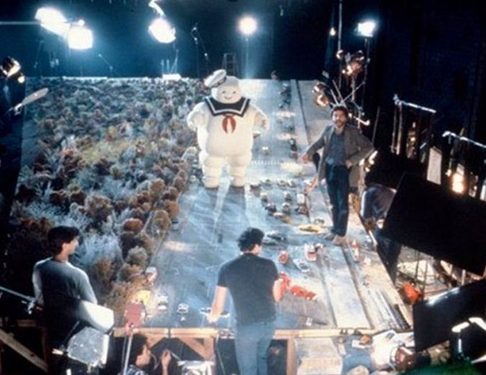 Unhinged behind-the-scenes photo showing a film crew and the Stay Puft Marshmallow Man on a detailed miniature set.
