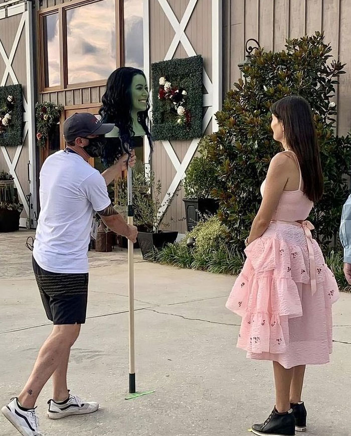 Behind-the-scenes photo showing a crew member holding a green-faced character head on a stick for a filming setup outdoors.