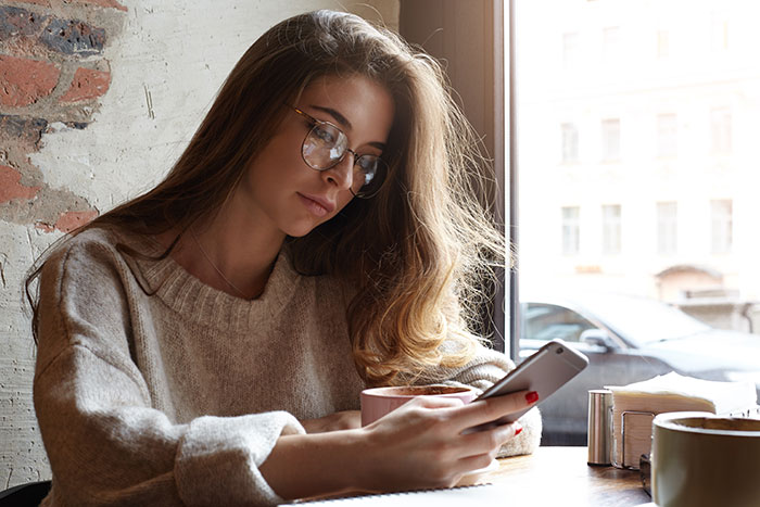 Young woman with glasses sitting by a window in a caf&eacute;, looking at her phone, reflecting on best friend no contact returns.