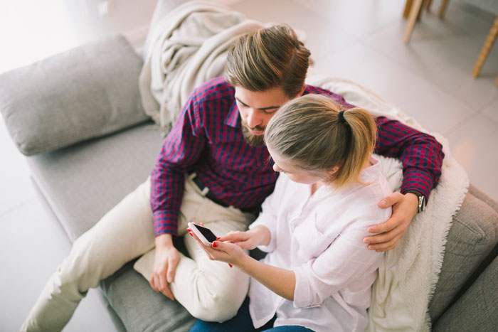 Couple sitting closely on a couch, woman showing phone to man, illustrating polyamorous woman and taken guy interaction.