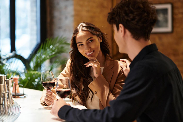 Polyamorous woman flirting with a taken guy at a bar while holding a glass of red wine in a cozy setting.