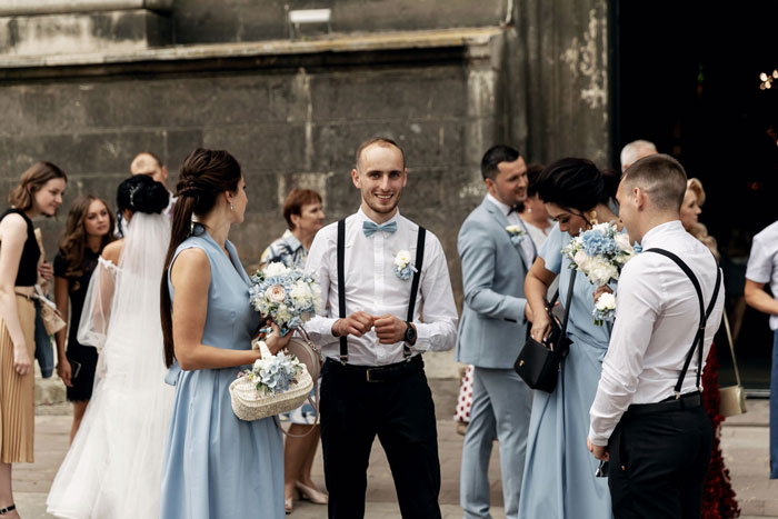 Polyamorous woman at a wedding socializing with a taken man while his girlfriend confronts her near a stone building.