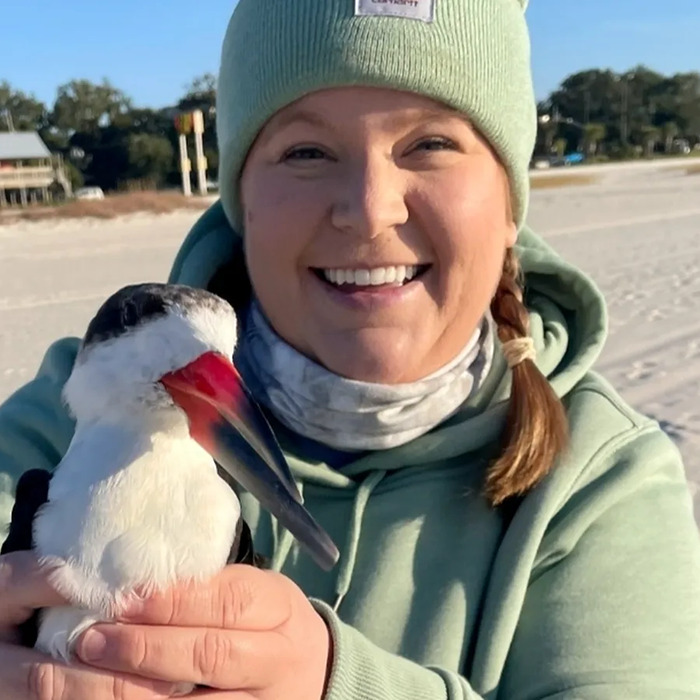 Smiling biologist wearing green hoodie and beanie holding a red-billed bird outdoors, related to biologist&rsquo;s life and lawsuit debate.