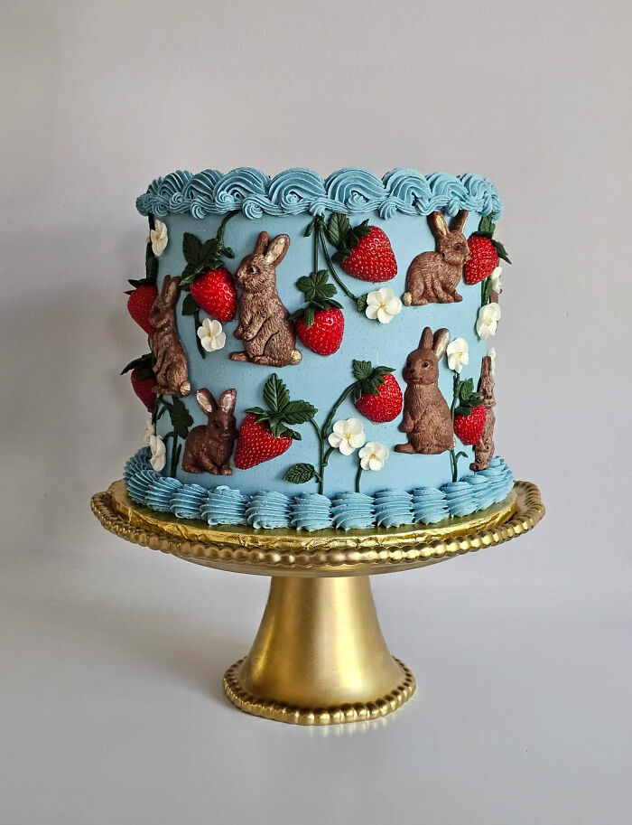 Blue frosted cake decorated with chocolate bunnies, strawberries, and white flowers on a gold cake stand, showcasing gorgeous baked goods.