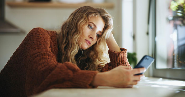 Young woman in a brown sweater looking thoughtful while using her phone during a family chat about birthday money.