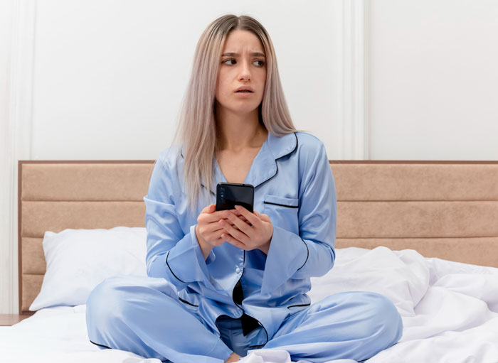 Woman in blue pajamas sitting on bed, looking worried while holding phone, hinting at boyfriend cheating on vacation.