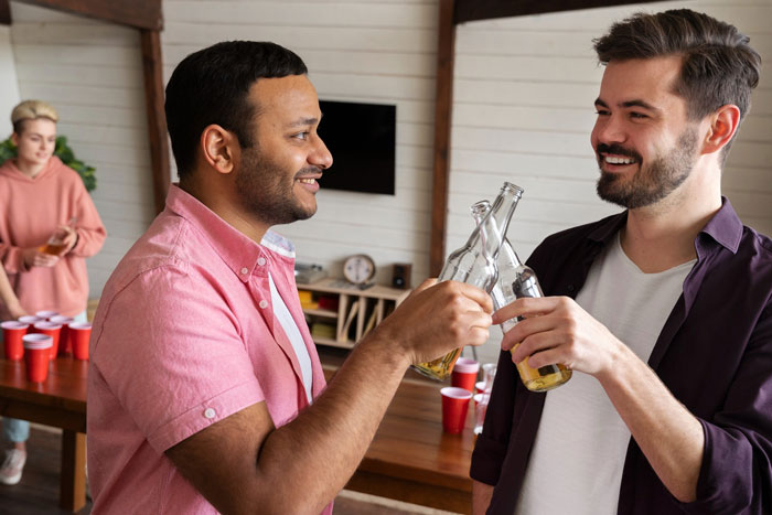 Two men clinking beer bottles at a casual party, while a woman stands in the background near a table with red cups.