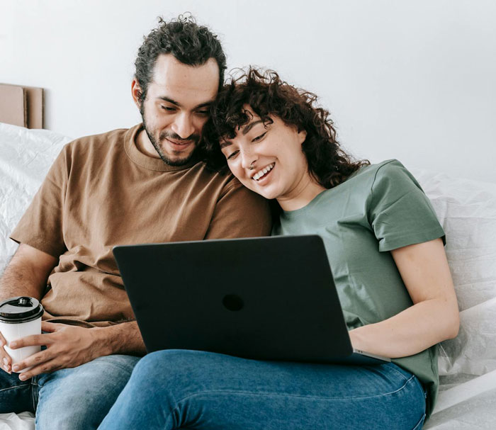 Couple sitting closely on a bed, smiling and looking at a laptop, illustrating a romantic trip with a female friend. Couple sitting closely on a bed, smiling and looking at a laptop, illustrating a romantic trip with a female friend.