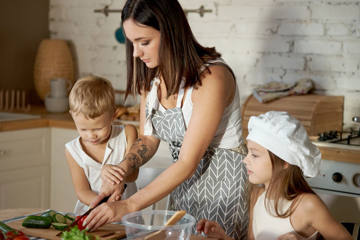 Woman acting as nanny with benefits, helping children prepare food in a cozy kitchen setting.