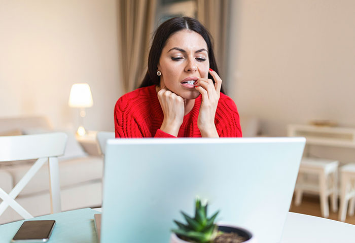 Worried woman in a red sweater biting nails while looking at laptop, reflecting bride mad cousin dress wedding stress.