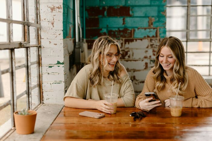 Two young women enjoying iced coffee and smiling while looking at a phone, illustrating culture shocks in Australia.