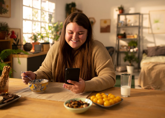 Young woman smiling at her phone while eating breakfast, reflecting on bro mocking sis over weight and diabetes.