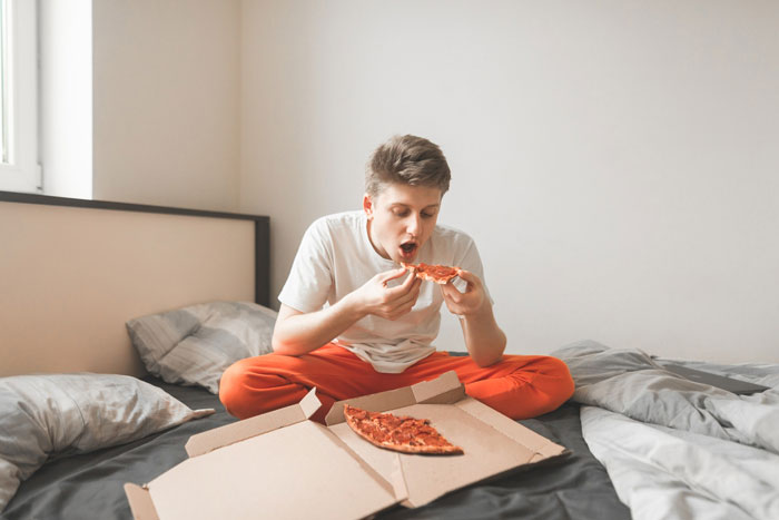 Teen boy eating pizza on bed with pizza box open, highlighting weight and diabetes themes in family dynamics.