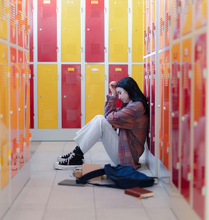 Teen girl sitting on school floor between lockers, distressed after relentless bullying over racist name at school.
