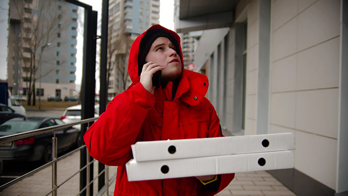 Delivery worker in a red jacket holding pizza boxes and talking on phone outside near apartment buildings representing business decisions.