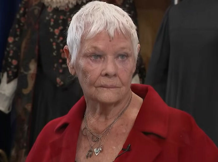Dame Judi Dench in a red coat, sharing a heartfelt health confession in an indoor setting with blurred background details.