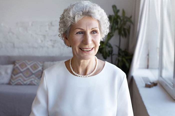 Elderly woman smiling indoors wearing a white blouse and pearl necklace near a bright window.